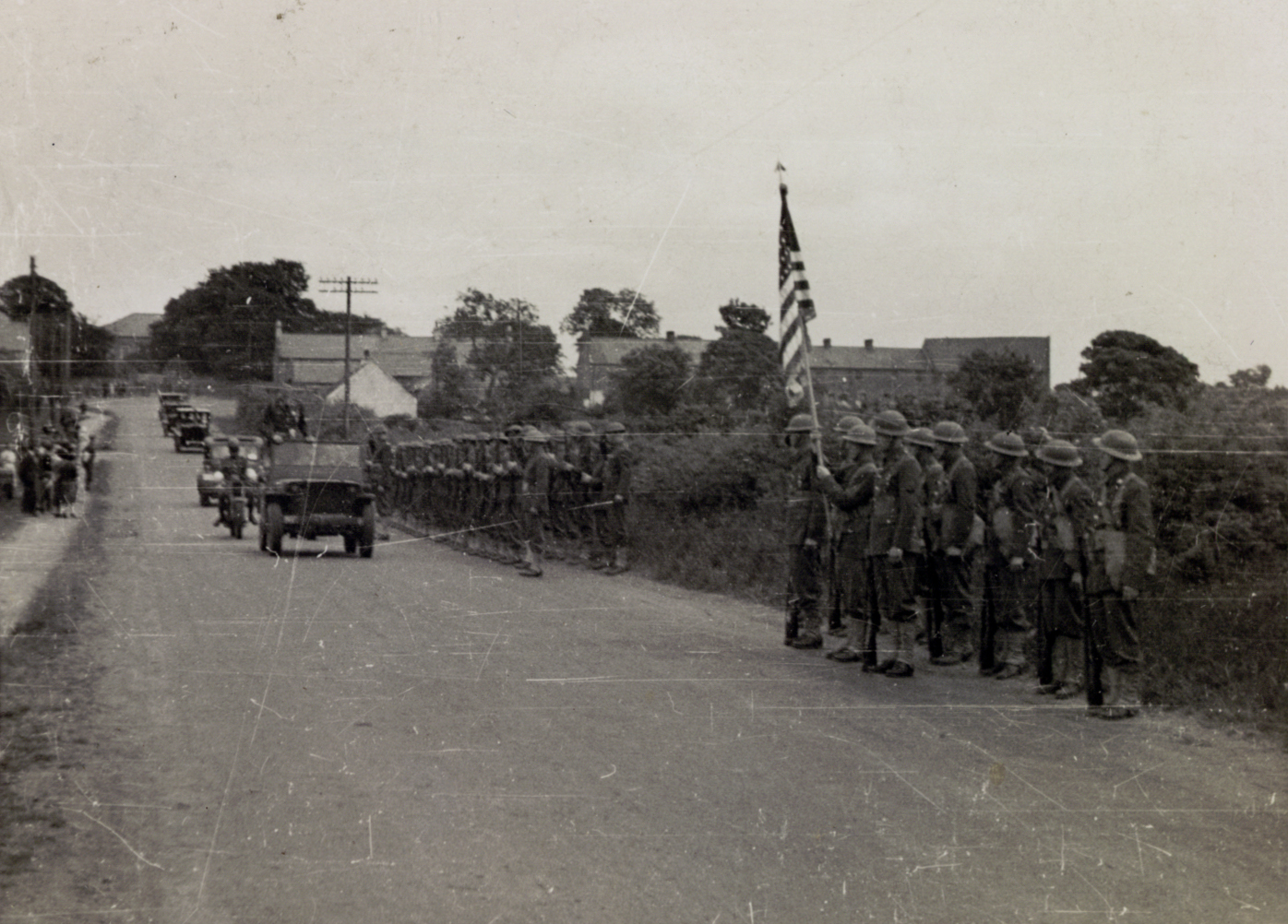 A wartime photograph of the procession through Clough: American soldiers at attention with the US flag along the road as a military motorcade passes.