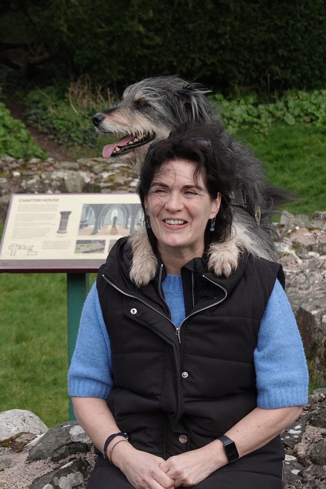 Lolly seated in the abbey grounds with a shaggy dog behind her, its paws draped over her shoulders.
