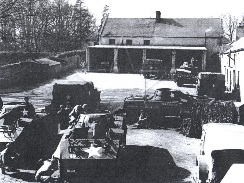 A wartime photograph of American armored vehicles parked in a courtyard, the same outbuildings behind and to the right.