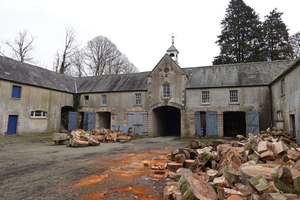 The stable courtyard, its clock tower and blue stable doors, piles of cut timber in the yard.